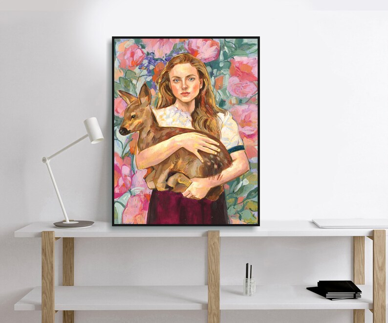 May include: A framed art print featuring a woman holding a fawn. The woman has long auburn hair and is wearing a white shirt with a dark red skirt. The background is a floral pattern with pink and green hues.
