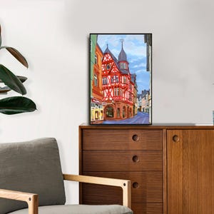 Pu&ograve; includere: Un dipinto ad acquerello incorniciato di una scena di strada europea con un edificio rosso e bianco. Il dipinto &egrave; esposto su un mobile in legno con una pianta e una tazza. Una sedia grigia e una grande pianta frondosa sono in primo piano.