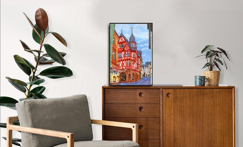Pu&ograve; includere: Un dipinto ad acquerello incorniciato di una scena di strada europea con un edificio rosso e bianco. Il dipinto &egrave; esposto su un mobile in legno con una pianta e una tazza. Una sedia grigia e una grande pianta frondosa sono in primo piano.