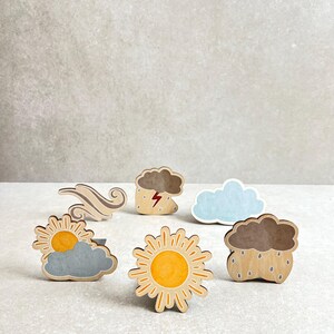 Könnte beinhalten: Holzstempel mit Wettersymbolen, darunter eine Sonne, eine Wolke mit Regen, eine Wolke mit Blitz, eine Wolke mit Wind und eine Wolke mit Sonne. Die Symbole sind in verschiedenen Farben bemalt, darunter Gelb, Blau, Grau und Braun.