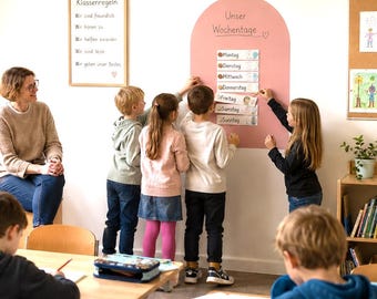 May include: A dusty rose arched wall decal labeled Unser Wochentage with removable day-of-the-week cards from Montag to Sonntag. The wall decal is displayed in a classroom setting with children interacting with the cards on the wall.