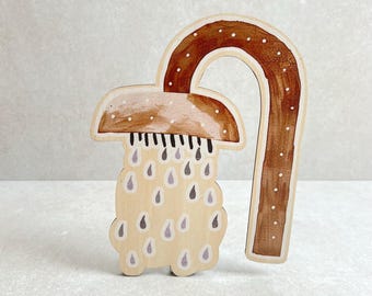 Puede incluir: Pieza decorativa de madera con forma de seta con un tallo curvo. El sombrero de la seta está pintado de marrón con puntos blancos, y el tallo es marrón. La seta tiene un cuerpo beige claro con detalles de gotas moradas.