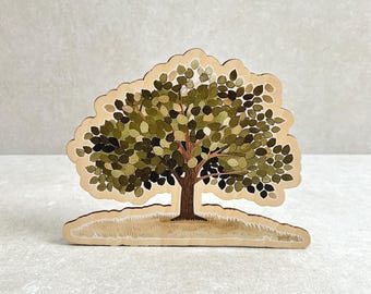 Puede incluir: Un recorte de árbol de madera con una ilustración detallada de un árbol con hojas verdes y marrones. El árbol está sobre una pequeña colina cubierta de hierba, todo dentro de un borde beige claro. El tronco es marrón y las hojas son de varios tonos de verde.