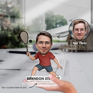 Op de afbeelding: Een gepersonaliseerde acrylkarikatuur van een squashspeler. De cartoonfiguur, gekleed in een rood shirt en een blauwe korte broek, houdt een racket en een bal vast. De basis van de figuur heeft de tekst "BRANDON STEWART SQUASH PLAYER". Een foto is inbegrepen.