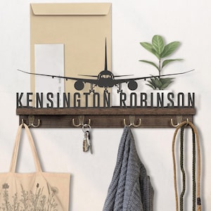 May include: A dark brown wooden coat rack with four brass hooks. A black metal airplane silhouette and the name "KENSINGTON ROBINSON" are above the hooks. A tote bag, keys, and a scarf hang from the hooks.
