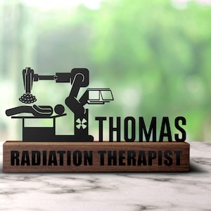 May include: A wooden plaque with a black metal cutout of a radiation therapy machine and a patient lying on a table. The plaque reads "Radiation Therapist" and "THOMAS".