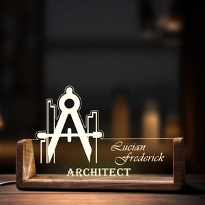 May include: A wooden desk nameplate with a clear acrylic insert. The insert features a compass and ruler design and the text "ARCHITECT". The nameplate also includes the name "Lucian Frederick".
