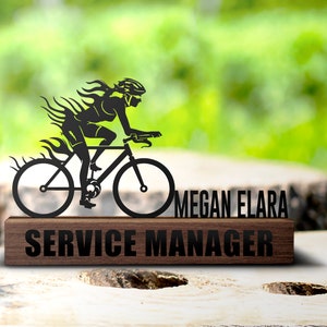 Könnte beinhalten: Schwarze Metallsilhouette einer Person, die ein Fahrrad mit Flammen dahinter fährt. Die Silhouette befindet sich auf einem Holzsockel mit dem Text "Megan Elara Service Manager".