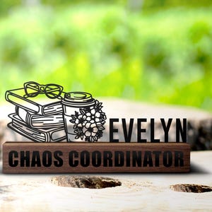 Może przedstawiać: Drewniany stojak na biurko z czarnym napisem "Evelyn Chaos Coordinator". Stojak przedstawia czarną sylwetkę stosu książek, filiżanki kawy z kwiatami i okularów.