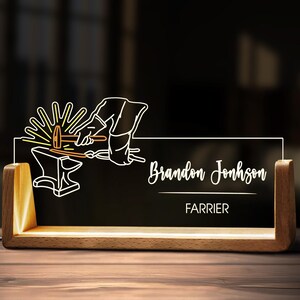 May include: A wooden desk lamp with a black acrylic panel. The panel features a white line drawing of a blacksmith at work, with the name "Brandon Johnson" and the word "Farrier" in white script. The lamp has a warm, inviting glow.