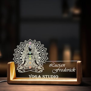 May include: A wooden desk sign with a lit acrylic panel featuring a white silhouette of a woman in a yoga pose with a mandala design and the text "Lucian Frederick Yoga Studio".