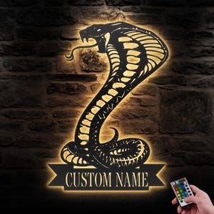 以下が含まれることがあります： 黒い金属製の壁飾りで、コブラと「CUSTOM NAME」と書かれたバナーが描かれています。暖色系の白色LEDライトで照らされており、リモコンでライトの色を変更できます。