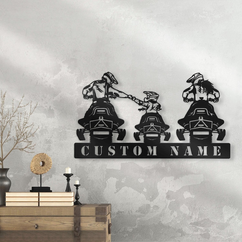 May include: Black metal wall art depicting three people riding snowmobiles. The snowmobiles are side-by-side and the people are holding onto each other. The art includes the text "CUSTOM NAME".