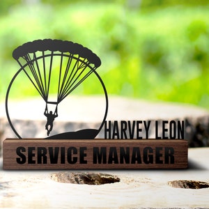 May include: A black metal desk plaque with a silhouette of a skydiver and a parachute. The plaque has a brown wooden base and the text "HARVEY LEON SERVICE MANAGER".