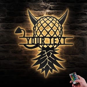 May include: A black metal pineapple with devil horns and a tail, with the text "YOUR TEXT" in the center. The pineapple is illuminated with warm white light. A remote control with multiple color options is shown in the bottom right corner.