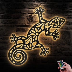 May include: Black metal gecko wall decor with a floral pattern. The gecko is illuminated with warm white light. A remote control is shown in the bottom right corner of the image.