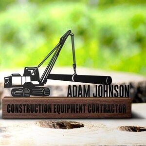 May include: Black metal silhouette of a crane lifting a log, mounted on a wooden plaque. The plaque reads "Adam Johnson Construction Equipment Contractor".