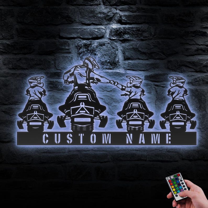 May include: A black metal silhouette of four snowmobilers with the text "CUSTOM NAME" below. The snowmobilers are riding in a line, with the first snowmobiler leading the way. The snowmobilers are all wearing helmets and goggles. The silhouette is illuminated with white light. A remote control with multiple colored buttons is visible in the bottom right corner of the image.