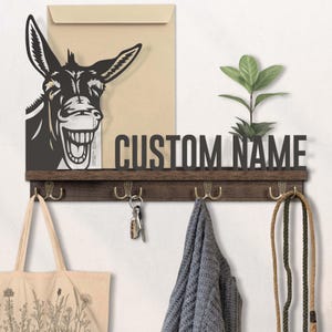 May include: A wooden wall-mounted key holder with a donkey silhouette and the words "CUSTOM NAME". It has four brass hooks, a small plant, and a tan tote bag hanging from it. A set of keys and a gray scarf are also hanging.