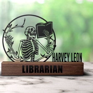 Puede incluir: Una silueta de metal negro de un esqueleto leyendo un libro con enredaderas que crecen a su alrededor. La silueta está montada sobre una base de madera con el texto "HARVEY LEON LIBRARIAN" grabado en ella.