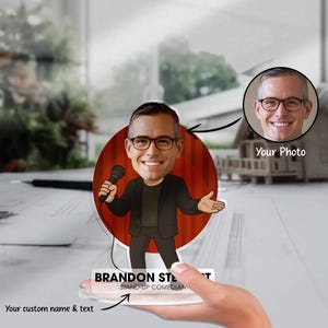 Puede incluir: Una caricatura personalizada de un comediante de stand-up con un nombre y texto personalizados. El dibujo animado presenta a un hombre sosteniendo un micrófono frente a un telón de fondo rojo. También se incluye una foto de la persona.