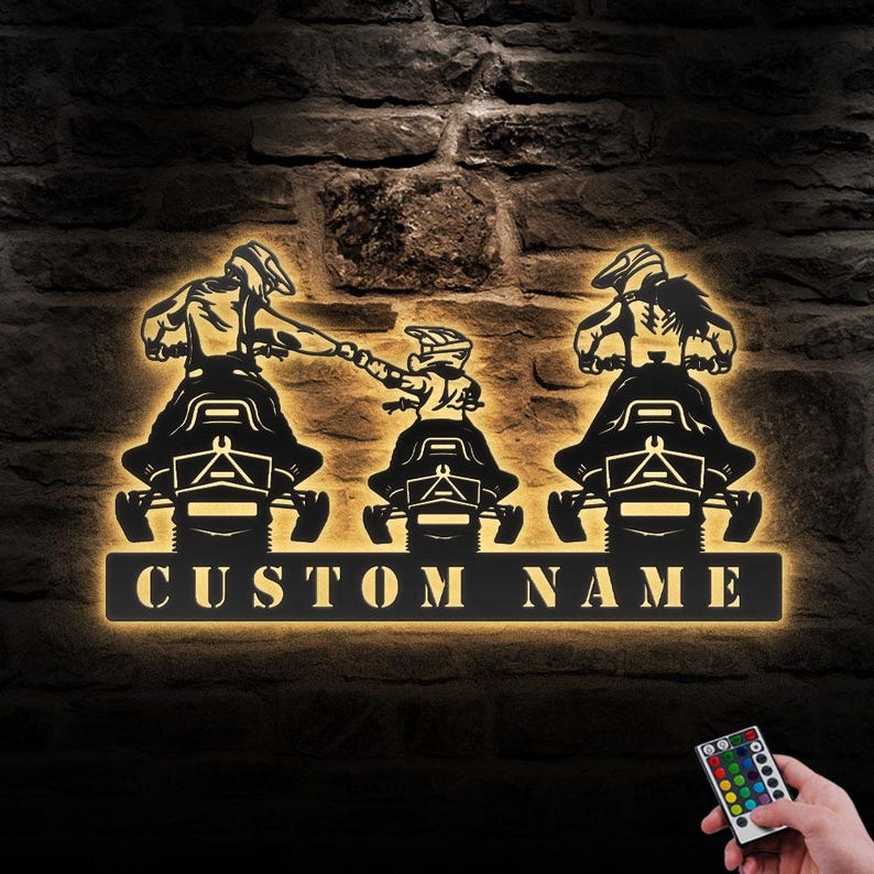 May include: A black metal silhouette of three people riding snowmobiles with the text "CUSTOM NAME" below. The silhouette is illuminated with warm white light and a remote control is visible in the bottom right corner.