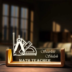 Op de afbeelding: Een houten bureau-naamplaatje met een transparante acryl-inzet. De inzet heeft een wit silhouet van een kompas, liniaal en geodriehoek. De tekst "MATH TEACHER" is onder de afbeelding gedrukt. De naam "Scarlett Shiloh" is in cursief boven de afbeelding gedrukt.