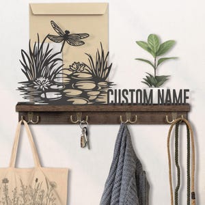 May include: A wooden wall-mounted shelf with hooks, a decorative dragonfly and pond scene, and the text "CUSTOM NAME". A small plant sits on the shelf. A tote bag, keys, and a scarf hang from the hooks.
