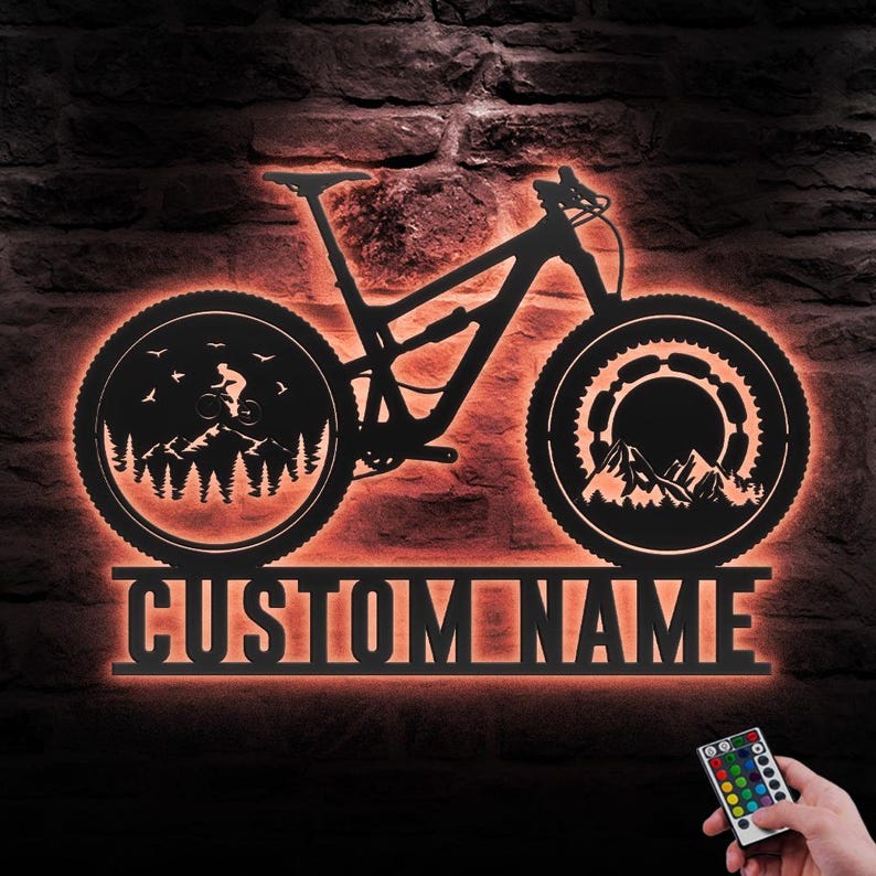 May include: Black metal mountain bike wall art with an orange LED glow. The bike silhouette features detailed wheels with mountain and forest scenes. The text "CUSTOM NAME" is displayed below the bike. A remote control is in the lower right corner.