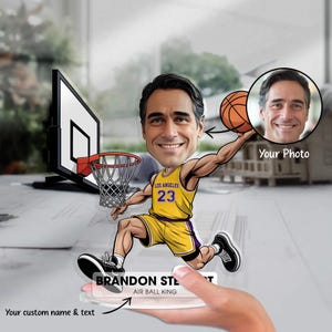 May include: A personalized basketball-themed acrylic figure featuring a cartoon caricature of a person in a yellow and purple jersey with the number 23. The figure is holding a basketball, with a basketball hoop in the background. The text "BRANDON STE" and "AIR BALL KING" is visible.