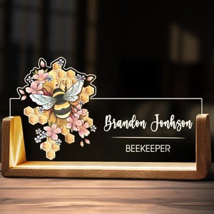 May include: A wooden desk nameplate with a black background. The nameplate features the name "Brandon Johnson" and the title "BEEKEEPER" in white script. A bee and floral design is on the left side of the nameplate.
