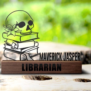 Puede incluir: Una placa de madera en blanco y negro con el texto "MAVERICK JASPER LIBRARIAN" y una calavera decorativa con una flor en la parte superior de una pila de libros.