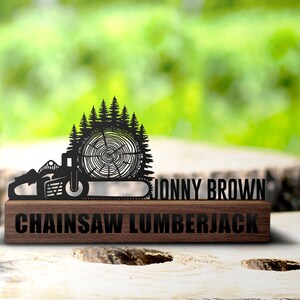 Könnte beinhalten: Schwarze Metallskulptur einer Kettensäge mit dem Namen "JONNY BROWN" und den Worten "CHAINSAW LUMBERJACK" auf einem Holzsockel. Die Skulptur zeigt eine Kettensäge, einen Baumstamm und eine Waldsilhouette.