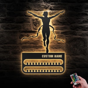 Puede incluir: Expositor de medallas de metal negro con una silueta de un corredor cruzando la línea de meta. El expositor tiene dos filas de ganchos para medallas y un espacio para un nombre personalizado. El expositor está iluminado con luces LED blancas cálidas y viene con un mando a distancia para cambiar el color de las luces.