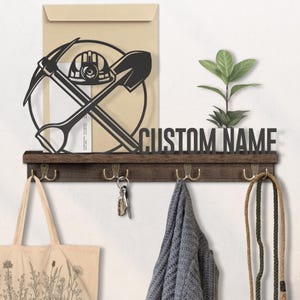 Puede incluir: Un llavero de pared de madera con ganchos, un estante y el texto "CUSTOM NAME". Presenta un diseño de metal negro de herramientas de minería y un casco. Una bolsa de mano, llaves y una bufanda cuelgan de los ganchos.