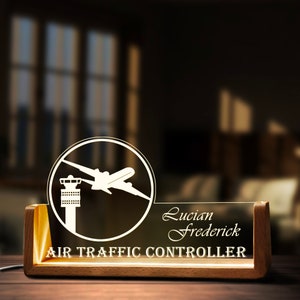 May include: A desk lamp with a wooden base and a clear acrylic panel. The panel displays a white graphic of an airplane and control tower, the name "Lucian Frederick", and the text "AIR TRAFFIC CONTROLLER".