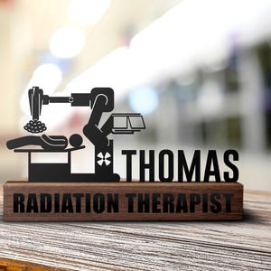 May include: A black metal silhouette of a radiation therapy machine with a patient lying on a table. The silhouette is mounted on a wooden plaque with the text "THOMAS RADIATION THERAPIST".