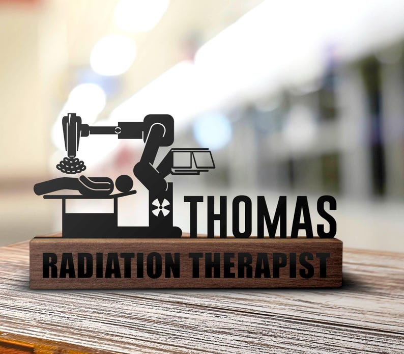 May include: A black metal silhouette of a radiation therapy machine with a patient lying on a table. The silhouette is mounted on a wooden plaque with the text "THOMAS RADIATION THERAPIST".