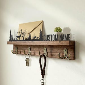 May include: A wooden key holder with four brass hooks. A black metal silhouette of a forest scene with deer and trees sits above the shelf. The text "CUSTOM NAME" is displayed on the shelf. A small succulent in a white pot and a brown envelope are also on the shelf.