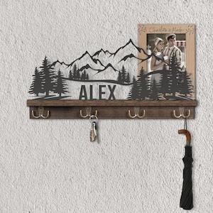 May include: A wooden key holder with a mountain and forest silhouette design. The name "ALEX" is cut out below the mountains. It includes a framed photo and four brass hooks. An umbrella and keys hang from the hooks.
