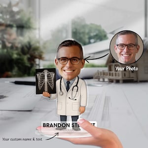 May include: A personalised doctor figurine holding an X-ray image. The figurine wears a white coat, glasses, and a stethoscope. The base of the figurine has the text "BRANDON STEWART RADIOLOGIST" and the words "Your custom name & text".