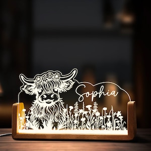 May include: A white acrylic night light with a wood base features a Highland cow with a floral crown and the name "Sophia" in a script font. The cow is surrounded by a field of flowers.