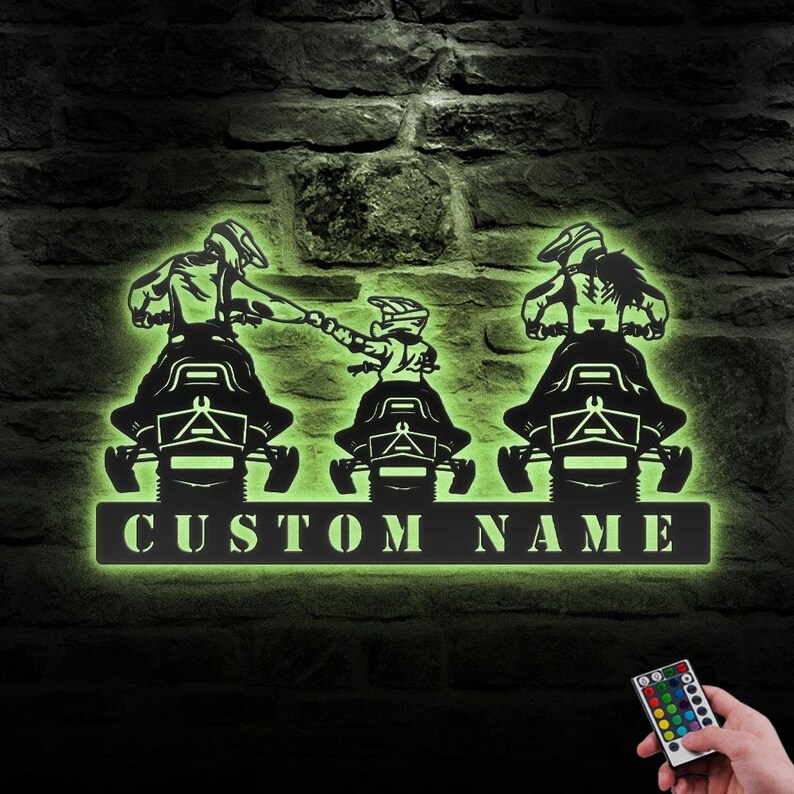 May include: A metal silhouette of three people riding snowmobiles with a customizable nameplate that reads "CUSTOM NAME". The silhouette is lit up in a green glow.