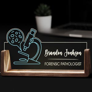 Puede incluir: Placa de escritorio iluminada con un diseño de microscopio azul. La placa dice "Brandon Jonhson, Forensic Pathologist" en escritura blanca. La base es de madera.