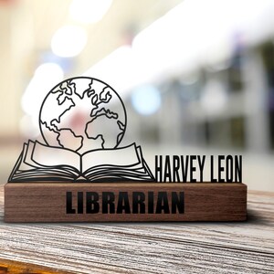 Puede incluir: Una placa de escritorio de madera con un contorno negro de un libro abierto y un globo en la parte superior. La placa dice "Harvey Leon Librarian".