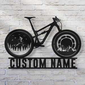 May include: Black metal mountain bike wall art with a detailed design. The wheels feature mountain and forest scenes. The text "CUSTOM NAME" is displayed below the bike. This decorative piece is mounted on a white brick wall.
