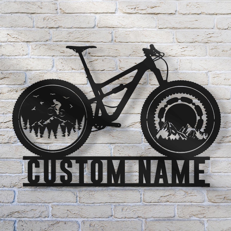May include: Black metal mountain bike wall art with a detailed design. The wheels feature mountain and forest scenes. The text "CUSTOM NAME" is displayed below the bike. This decorative piece is mounted on a white brick wall.