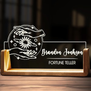 May include: A lighted desk nameplate with a wooden base. The black acrylic panel displays the name "Brandon Johnson" and the title "Fortune Teller" in white script. A white graphic of hands, a sun, and stars is above the name.