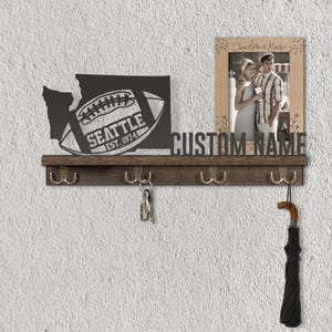 May include: A wooden wall-mounted key holder with a dark finish. It features a metal cutout of the Washington state outline with a football and the text "SEATTLE EST. 1974". Includes a photo frame and hooks for keys and an umbrella.
