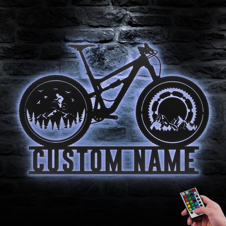 May include: Black metal mountain bike wall art with a blue LED backlight. The bike silhouette features a rider, trees, mountains, and the text "CUSTOM NAME". A remote control is visible in the bottom right corner.